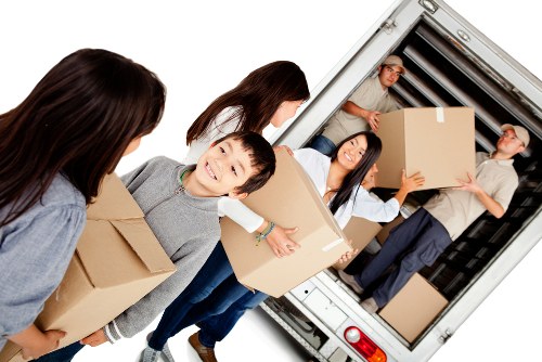 A group of five individuals, including children and adults, are engaged in loading a large removals van parked in a residential area. The scene is set in front of a house with a visible driveway, with the van positioned nearby. The individuals are carrying cardboard boxes of various sizes; some are plain brown, and one person in the background is holding a box with a smile, indicating a positive atmosphere. The boxes include a mix of plain and taped packaging. One woman is holding a large box close to her chest, while a young boy in the foreground is about to hand over or receive a box from another person. The interior of the van shows more boxes stacked neatly, with some personal belongings possibly wrapped or protected by blankets or padding. The people are dressed in casual clothing suitable for moving, and the natural lighting suggests daytime. This scene reflects professional house removal services offered by Westminster Man with Van in the London area, subtly supporting local SEO for house removals and related logistics in Westminster or central London postcode regions.