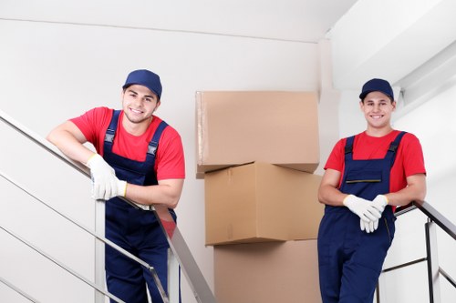Two professional removal men from Westminster Man with Van, dressed in red shirts, navy overalls, and matching caps, are standing on a staircase landing inside a residential property in Westminster. They are smiling and wearing white gloves while posing for the camera. Behind them, there is a stack of three large cardboard moving boxes, with two on top and one below, all sealed and ready for transport. The setting features a white-painted interior wall and a metal railing along the staircase, with natural lighting illuminating the scene. The environment suggests an organized, interior house removal process in a London postcode area, emphasizing careful handling of household items. The removal specialists appear prepared to load or unload belongings, illustrating professional house relocation services offered by Westminster Man with Van. The image communicates readiness and professionalism in facilitating safe and efficient removals for clients in central Westminster or nearby areas.