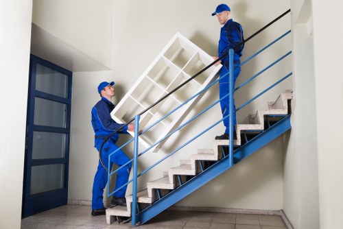 Two professional movers from Westminster Man with Van are seen carrying a large, white bookshelf with multiple compartments up a staircase inside a modern residential building. The staircase has wooden steps with a black tread pattern and is enclosed by a blue metal railing, which the movers are holding onto for support. The background reveals a plain white wall and an open door with glass panels on the left, leading to another room. The movers are dressed in matching blue uniforms and caps, and are working carefully to navigate the bookshelf over the stairs. Natural light illuminates the area, creating a clear and professional scene typical of house removals in Westminster and surrounding areas. The environment is tidy and functional, with the interior appearing minimalist, designed to facilitate efficient moving activities within the property.