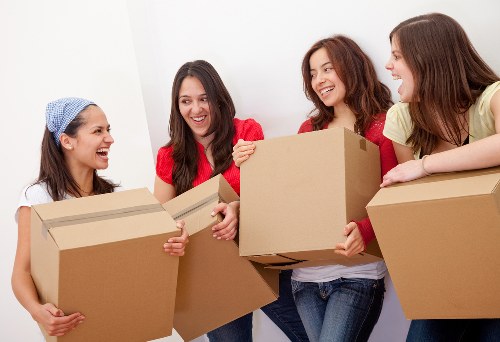 Four women indoors, standing close together, engaged in a cheerful interaction while holding and passing large cardboard boxes. The woman on the far left, wearing a light blue headscarf and white top, holds a box with both hands. The second woman from the left, dressed in red, smiles as she offers a box towards the group. The third woman, also smiling, has shoulder-length brown hair and wears a white top with a red cardigan, holding a box in front of her. The woman on the far right, with dark brown hair and wearing a yellow top, rests her hands on a box placed on a surface, possibly a table or ledge. The background features a plain white wall, indicative of a domestic or residential setting typical of house removals or relocation scenes, subtly reflecting a UK environment. The natural lighting enhances the friendly atmosphere of a house moving process, with the women appearing to plan or prepare for a move, aligned with services offered by Westminster Man with Van in the local Westminster or nearby postcode areas.