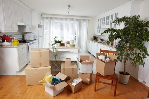 A bright, spacious kitchen with white cabinets and countertops, illuminated by natural light from large glass doors with white vertical blinds. The floor is made of light wooden planks. In the center of the room, numerous cardboard boxes, some open and partially filled with household items such as paper, plastic containers, and packing materials, are scattered across the floor. A small stack of boxes is placed on a dark patterned bag, and additional boxes are positioned on a wooden chair situated on the right side of the image. There is a large potted plant with lush green foliage on the far right, next to the chair. On the countertops, various kitchen appliances, utensils, and small decorative items are visible, suggesting a house relocation in progress. The scene depicts an active packing or unpacking process, typical of a house removals service in Westminster or nearby areas, with the environment clean, well-lit, and organized for an upcoming move.
