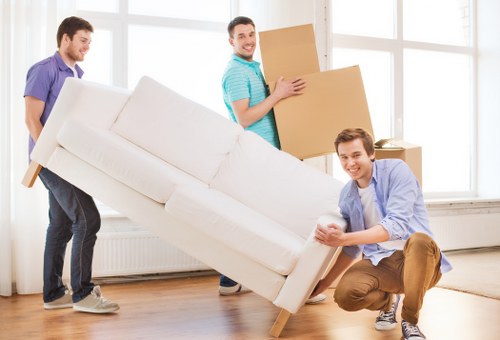 Three young men with light skin and casual clothing are engaged in a house removals task inside a bright, spacious room with large windows. One individual, wearing a blue T-shirt, is smiling and holding a large cardboard box, preparing to move it across the room. Another man, dressed in a purple T-shirt, is helping to carry a light-colored fabric-upholstered sofa, which appears to be mid-motion and slightly tilted. The third person, wearing a light blue shirt and brown trousers, is crouching and steadying the sofa, ensuring it does not tip over. The room features a wooden floor, a white radiator beneath the windows, and minimal furnishings, emphasizing the focus on moving household items such as the sofa and cardboard boxes, typical of house removals in the Westminster or central London area. The scene is well-lit with natural sunlight, capturing a moment of teamwork during a residential relocation by Westminster Man with Van within the London postcode area.
