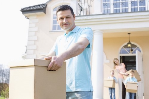 A professional mover from Westminster Man with Van stands outside a residential property, holding a cardboard box in front of him. The house features a cream-colored facade with decorative white trim and a large window with white framing on the upper level. In the background, a woman and two children are visible, each carrying similar cardboard boxes, and standing on the paved pathway leading to the house's entrance. The porch is supported by white columns, and a black outdoor light fixture hangs from the ceiling. The scene is set during daylight hours with natural lighting, capturing the process of house removals and the careful handling of household items such as boxes. The setting is a typical suburban street in Westminster, with visible elements of a well-maintained home ready for relocation, emphasizing professional residential moving services.