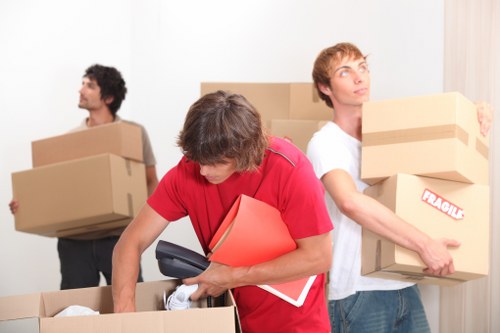 Three men involved in a house move inside a bright, minimally decorated residential room. The man in the foreground, wearing a red shirt, is leaning over a cardboard box while holding a black cordless drill, a red folder, and some papers, appearing to be organizing packing or preparing for loading. Behind him, another man with light brown hair and a white t-shirt is carrying a medium-sized cardboard box labeled 'Fragile', holding it securely with both hands, and looking slightly upward. To the left, a third man with dark, curly hair in a gray t-shirt is standing with a large cardboard box in his arms, facing sideways. The background features a plain white wall, and the scene is illuminated with natural daylight, emphasizing the clarity of the cardboard boxes, which vary in size and are packed with household items typical of a home removal, such as boxes, documents, and packing materials. The environment suggests an organized move within a residential property, with a focus on professional house removals, consistent with services offered by Westminster Man with Van in the Westminster or nearby London area.