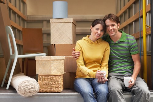 A young couple sits on the floor of a house storage area or moving van, surrounded by packed cardboard boxes, some with textured surfaces and brown paper finishes, stacked in front of a bookshelf or shelving unit. The woman wears a bright yellow hoodie and blue jeans, holding a clear glass mug, while the man, dressed in a green and purple striped polo shirt and grey pants, holds a white mug. Behind them, there are additional boxes, a rolled-up white carpet, and a light blue container placed on top of a large beige storage box. The background features wooden walls and shelves, indicating an indoor space used for storage during a house move in Westminster or nearby. Natural indoor lighting illuminates the scene, reflecting the calm and organized atmosphere of the home relocation process. Westminster Man with Van’s professional removals services are suggested by the context, with the couple appearing relaxed in the midst of packing or settling during their move in the London area, possibly around SW1 or SW7 postcode zones.