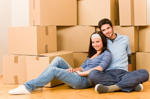 A young couple sits on a wooden floor inside a residential space, surrounded by numerous brown cardboard moving boxes of varying sizes, some stacked against the wall and others arranged around them. The woman, wearing a white headband and casual blue jeans with a matching blue top, is comfortably seated with her legs extended and crossed at the ankles, smiling gently at the camera. The man, dressed in a light blue shirt and dark jeans, sits behind her with his legs stretched out, leaning slightly on his left arm, also smiling. The background features a plain wall and a doorway leading to another room, with the scene illuminated by soft, natural lighting. The boxes, some sealed with packing tape, suggest an ongoing house move typical of residential relocation services such as those offered by Westminster Man with Van, serving local areas including Westminster and nearby London districts. The image emphasizes packing and moving preparation in a home environment, relevant to house removals and relocating services.