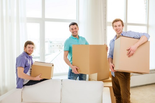 The image depicts three men in a bright, modern residential living room with large windows allowing natural light to fill the space. They are engaged in moving and carrying cardboard boxes, which are of various sizes, some sealed and some open, revealing household items inside. The men are dressed casually in shirts and trousers, with one wearing a purple shirt, another in a blue and white striped shirt, and the third in a light blue shirt. They are positioned around a white sofa, with one man seated on the armrest holding a medium-sized box, while the other two stand on either side of the sofa, each holding a larger box. The environment suggests an organized move within a home, possibly in Westminster or a nearby area, as supported by the context of a house removals service. The background includes the expansive windows with white frames, and the scene’s neutral tones of furniture and boxes contrast with the bright daylight, emphasizing the active loading or packing process typical of professional house removals in London or the surrounding region. The overall setting portrays a professional, orderly move carried out by Westminster Man with Van, highlighting their removals service capabilities.