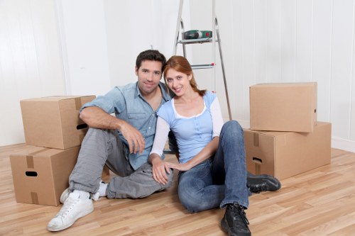 A young man and woman sit on a light wooden floor in a bright, modern interior adjacent to a set of cardboard boxes, indicating a house move. The man, dressed in a light blue shirt and grey jeans, has dark hair and a slight smile, while the woman, with red hair, wears a blue top and jeans, leaning slightly towards him. Behind them, there is a silver stepladder with a few small tools or items placed on its steps, set against a white paneled wall. Several large cardboard moving boxes are stacked around them, some closed and others open, prepared for packing or unpacking. The environment suggests a residential setting, likely within a house or apartment in Westminster or nearby, with a clean, uncluttered space ready for a house removals service. The scene captures a moment of preparation or transition typical of domestic relocations undertaken by a professional removal company like Westminster Man with Van, with natural lighting highlighting the wooden flooring and neutral backdrop.