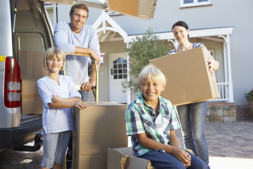 A family of four is engaged in a house removal process outside a residential property in Westminster, London. The father, wearing a light blue shirt, stands next to an open removal van, smiling as he leans on a large cardboard box positioned on the van's cargo area. The mother, dressed in a striped top, holds a medium-sized cardboard box, ready for loading or unloading. Two children, a girl with blonde hair and a boy with blonde hair and a plaid shirt, are seated on stacked cardboard boxes on the driveway, smiling at the camera. The scene is set on a paved driveway in a suburban neighbourhood, with a light blue house featuring a white porch and windows visible in the background. The van appears to be parked on a residential street, and the overall lighting suggests a bright, clear day. The family and items are arranged in front of the vehicle, indicating an active moving or relocation effort, consistent with local house removal services in Westminster.