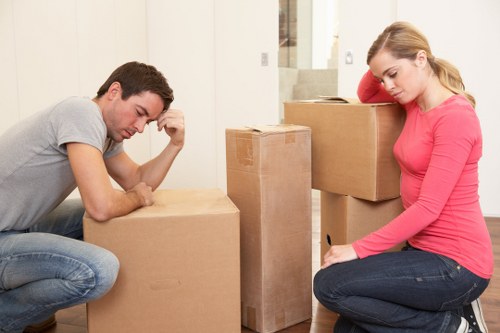 A young man and a young woman are in a brightly lit room with white walls, preparing for a house move in Westminster. The man, wearing a grey t-shirt and jeans, is kneeling on the floor, leaning forward with his elbow resting on a large, sealed cardboard box, appearing contemplative. The woman, dressed in a pink long-sleeve top and dark jeans, is seated on the floor beside a stack of moving boxes, with her head resting on her hand and her eyes closed, suggesting a moment of rest or fatigue. Several filled and unopened cardboard boxes of various sizes are arranged on a wooden floor, some of which are stacked and others placed individually. The backdrop features a staircase and a hallway indicating an interior residential space. The cardboard boxes have a natural brown kraft finish and are closed, ready for transportation. This scene exemplifies typical household packing activities associated with professional removals services offered by Westminster Man with Van, serving areas around Westminster and central London, with a focus on efficient and careful packing of personal belongings in preparation for a move within the UK.