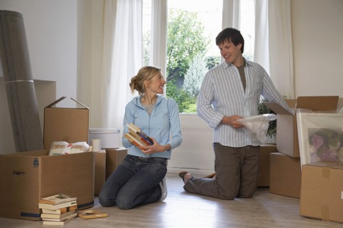 In a well-lit room with large windows revealing greenery outside, two individuals are engaged in a house removals process. The woman, with blonde hair tied back and dressed in a light blue shirt and dark jeans, is kneeling on the wooden floor and holding a stack of books and a small box, appearing to discuss or prepare for packing. The man, wearing a striped shirt and grey trousers, is kneeling beside a large cardboard box filled with household items, including a plastic-wrapped object and smaller boxes. Surrounding them are several open cardboard boxes of varying sizes, some filled with books, and others containing household belongings. The scene captures the process of packing up a residential space, typical of a professional removals service in Westminster or surrounding areas, with natural daylight illuminating the activity. The background features neutral curtains and a glimpse of outdoor greenery, creating a calm, functional environment suited for a house moving operation.