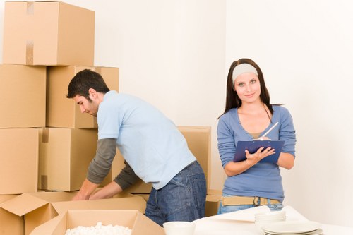 In a domestic setting with plain white walls, a man with dark hair and a beard is seen packing or unpacking cardboard moving boxes, some of which are stacked in the background, while others are open and partially filled with packing material; he is wearing a light blue t-shirt with a grey long-sleeve shirt underneath and jeans. Next to him, a woman with long dark hair, wearing a pink headband, a blue long-sleeve top, and light-colored trousers, holds a clipboard and pen, possibly coordinating the packing process. The scene takes place in a room likely part of a house in Westminster or nearby in London, with soft, natural lighting illuminating the area, and a white table visible in the foreground. The image relates to house removals and relocation services offered by Westminster Man with Van, showcasing packing activities typical of a move, with a focus on household items such as boxes and packing supplies.