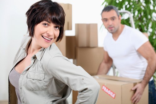 A woman with short dark hair and a light grey jacket smiling directly at the camera, standing in a residential interior with a bright atmosphere. Behind her, a man with short dark hair and a white t-shirt is slightly out of focus, holding a cardboard box with a red and white label. The background includes several stacked cardboard boxes of various sizes, some on the floor and others on a trolley or dolly, indicating an ongoing house removal or relocation process. The environment suggests a home or flat with a modern interior, featuring neutral tones and a large green plant visible behind the man. The scene captures the preparation or packing stage of a house removal service, delivered by Westminster Man with Van, highlighting professional household moving activities typical of London suburbs or nearby areas. Visibility includes different household items such as moving boxes, the woman's casual clothing, and the man's casual attire, with natural lighting illuminating the interior spaces clearly.