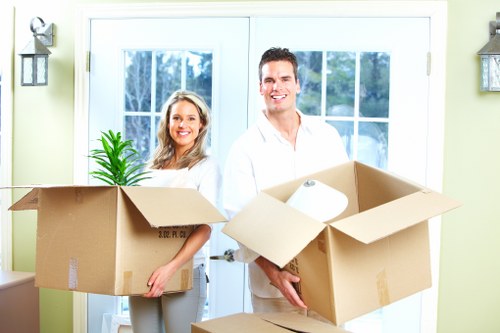 A smiling man and woman standing inside a bright residential room near a large glass door, holding open cardboard moving boxes. The woman, positioned on the left, has long blonde hair and is wearing a light-colored blouse and trousers, carrying a box that contains a small potted plant with green foliage. The man, on the right, has short dark hair and is dressed in a white shirt and light-colored trousers, holding an open box with a white rolled-up item inside. Behind them, the room features a pale yellow wall with two wall-mounted outdoor lanterns, and the large glass door provides natural light and a view of a garden or outdoor space. The scene captures a moment of packing or preparing for a house move, indicative of the services offered by Westminster Man with Van, a removals company operating in the Westminster area. The environment is clean, well-lit, and orderly, emphasizing a professional and efficient moving process within a typical UK home setting.