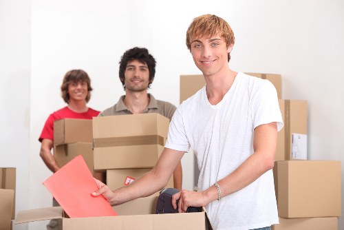 Three young individuals in a bright, spacious interior surrounded by cardboard boxes are preparing for a house move; a smiling man in a white t-shirt at the forefront is placing a red envelope into an open cardboard box, with a silver bracelet on his wrist. Behind him, a woman with shoulder-length brown hair wearing a red top holds a box, and a man with dark hair and a beard, dressed in a casual beige shirt, also carries a box. The scene suggests a professional house removal by Westminster Man with Van, with a focus on the careful packing and handling of household items, such as cardboard boxes and envelopes, set against a plain white background typical of a residential interior or a moving depot, subtly indicating local services in Westminster, London, as part of a removals process. The lighting is natural and evenly distributed, emphasizing the clean and organized environment suitable for a house removals team engaged in packing or transporting household belongings.