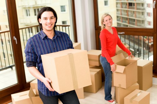 Two individuals are inside a residential property, preparing for a house removal in Westminster. The man in the foreground, with dark hair and wearing a blue striped shirt, is holding a medium-sized cardboard box sealed with brown packing tape, displaying a neutral yet focused expression. Behind him, a woman with blonde hair dressed in a red long-sleeve top and jeans is stacking and organizing several cardboard boxes of varying sizes, some open and some sealed. The boxes appear to contain personal belongings or household items and are arranged on a light wooden floor near large windows that let in natural daylight. The windows provide a view of a residential street with multi-story buildings and balconies, indicating an urban London setting. A small balcony with metal railings can be seen through the windows. The interior space has a modern appearance with wooden window frames and a bright, tidy atmosphere, typical of a professional house removals service operating in the Westminster area. Westminstermanwithvan.co.uk logo and website are associated with this scene, focusing on home relocation tasks in central London neighborhoods.