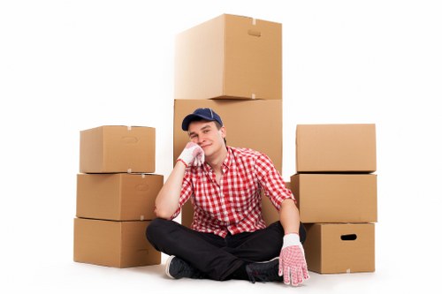 A young man wearing a blue cap, red and white checkered shirt, black trousers, and pink work gloves is sitting on the floor of a plain white background, surrounded by neatly stacked cardboard moving boxes of varying sizes. He is resting his chin on his right hand, with a slight smile, while his left hand rests on the floor. The boxes are positioned behind, to the left, and to the right of him, some with cut-out handles visible on their sides. There are no other objects or environmental details in the scene, emphasizing a simple and professional setup likely representing house removals. The plain background and positioning of the boxes suggest an indoor setting, possibly a residential property in Westminster or nearby London areas, consistent with the services of Westminster Man with Van. The arrangement highlights the process of packing and relocation, aligning with house removals and moving services available in the local postcode areas.